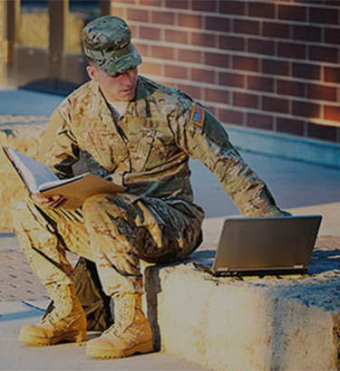 man in a military uniform sitting with a book in hand and laptop to his side